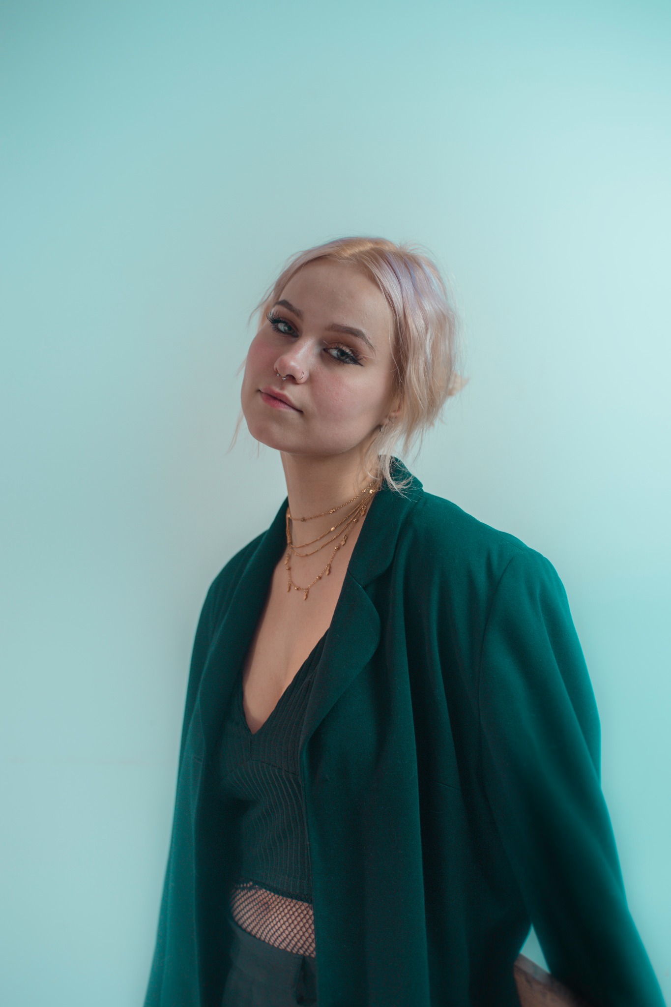 Portret młodej kobiety z blond włosami z fioletowymi pasemkami, ubranej w czarny top i zieloną marynarkę, pozującej na tle jasnoniebieskiej ściany, z trzema złotymi łańcuszkami na szyi...