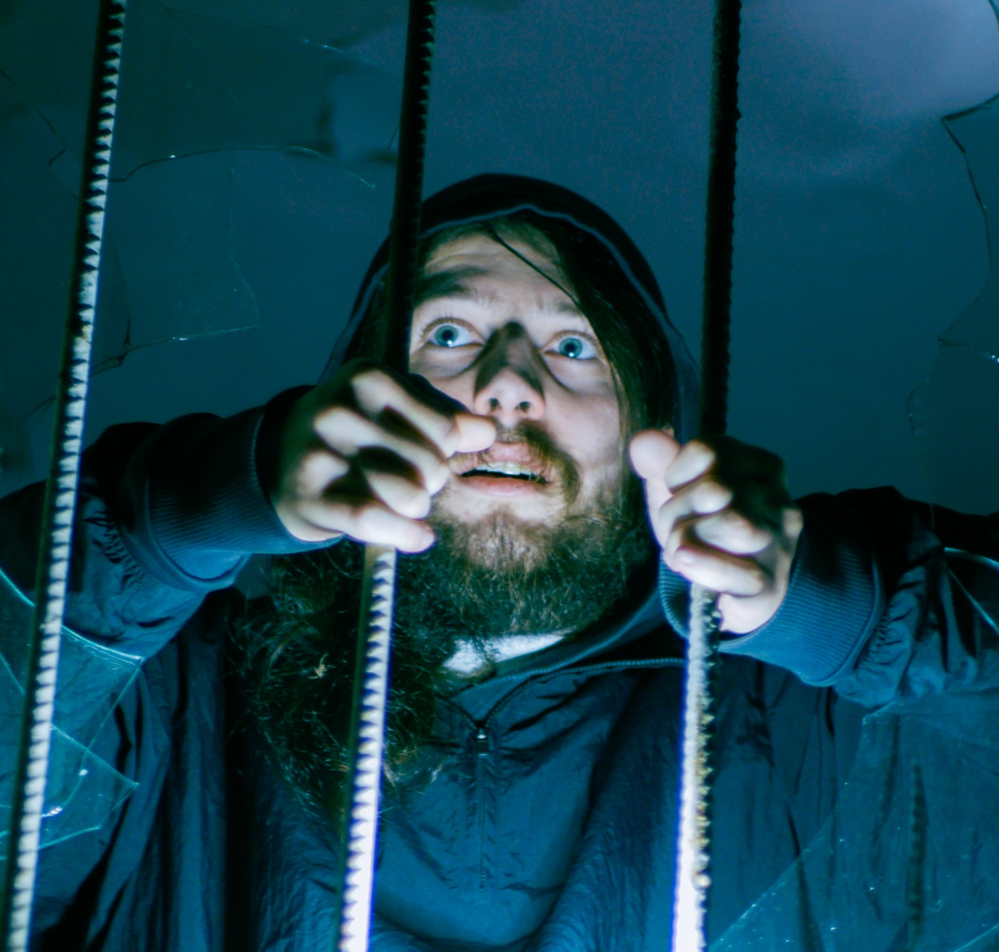 Mężczyzna z brodą i w kapturze, z przerażeniem w oczach, trzyma się krat zza potłuczonego szkła, oświetlony od dołu niebieskim światłem.