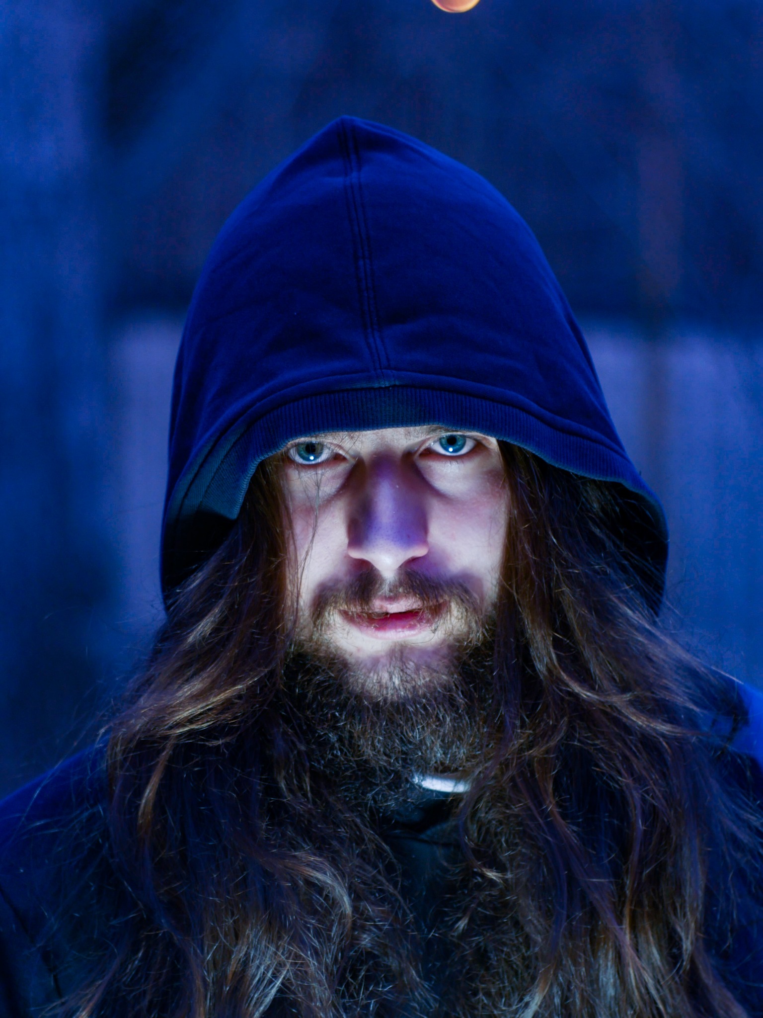 Portret mężczyzny z długimi włosami i brodą w niebieskim oświetleniu, ubrany w bluzę z kapturem, z intensywnym spojrzeniem skierowanym wprost.