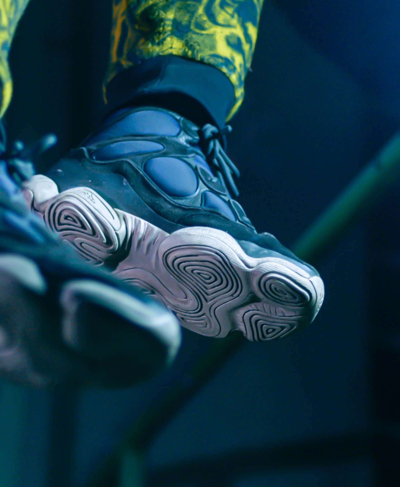 Zbliżenie na podeszwę i fragment cholewki sportowego buta w odcieniach granatu i bieli, z widoczną teksturą podeszwy przypominającą labirynt, oraz fragment spodni w żółto-czarny wzór na górze kadru.