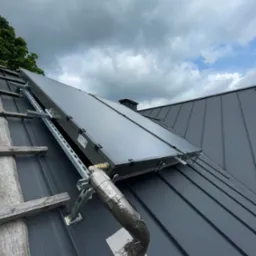 Dwa panele solarne zamontowane na metalowym dachu z blachy na systemie wsporników, widoczne drewniane elementy rusztowania i fragment instalacji z izolacją termiczną.