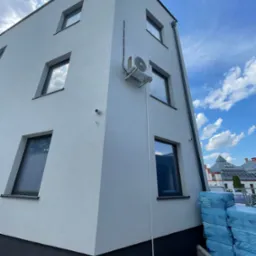 Zewnętrzna jednostka klimatyzacji LG zamontowana na elewacji nowoczesnego budynku z widocznymi oknami i rurami instalacyjnymi, w tle błękitne niebo z chmurami i elementy sąsiednich budynków.