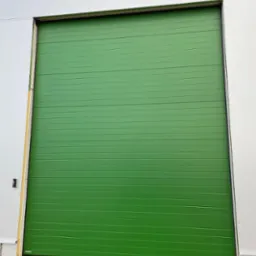 Częściowo otwarta, segmentowa brama w kolorze zielonym, zamontowana w białej hali przemysłowej, widoczna kostka brukowa na dole.