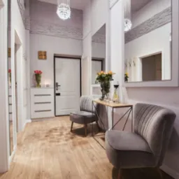 Jasne wnętrze przedpokoju z drewnianą podłogą, szarymi krzesłami i lustrami na ścianach, oświetlone kryształowym żyrandolem.