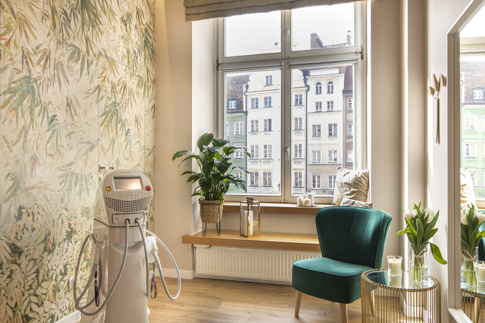 Jasne wnętrze gabinetu z aparaturą do zabiegów, fotel w odcieniach zieleni, okno z widokiem na kamienice, ściana w roślinne wzory.