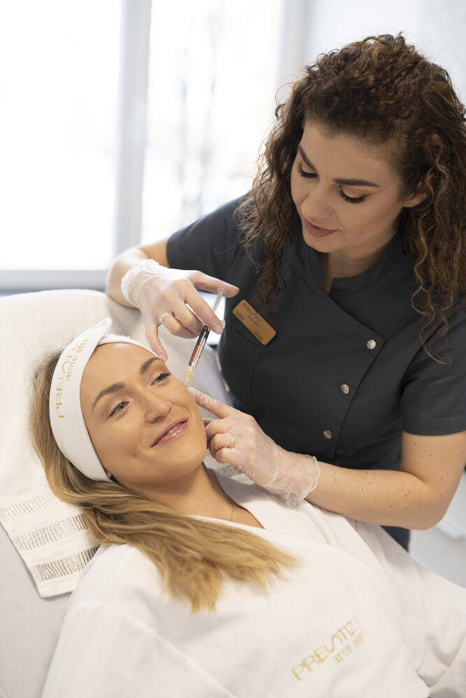 Klientka leżąca na leżance w białym szlafroku i opasce na włosach, podczas zabiegu z użyciem strzykawki, wykonywanego przez kosmetologa w rękawiczkach.