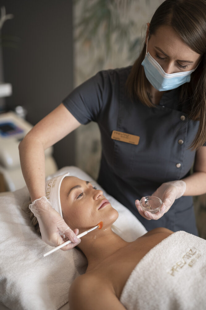 Kosmetolog w rękawiczkach i masce aplikuje pędzlem preparat na twarz klientki leżącej na fotelu zabiegowym, w tle widoczny sprzęt kosmetyczny.