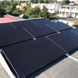 Panele słoneczne zamontowane na płaskim dachu budynku mieszkalnego, widoczne elementy konstrukcji wsporczej, w tle zabudowa miejska i zieleń.