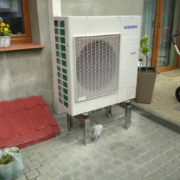 Zewnętrzna jednostka klimatyzacyjna Samsung zamontowana na metalowej konstrukcji, widoczna na tle szarej kostki brukowej i fragmentu czerwonej blachy dachowej.