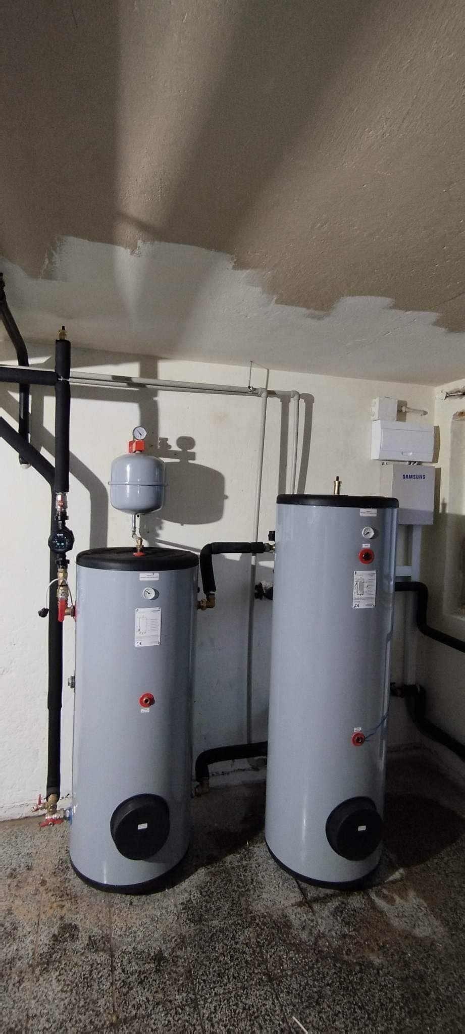 Dwa szare zbiorniki akumulacyjne ciepłej wody w piwnicy, z widocznymi rurami, manometrami i osprzętem; obok biała skrzynka Samsung.
