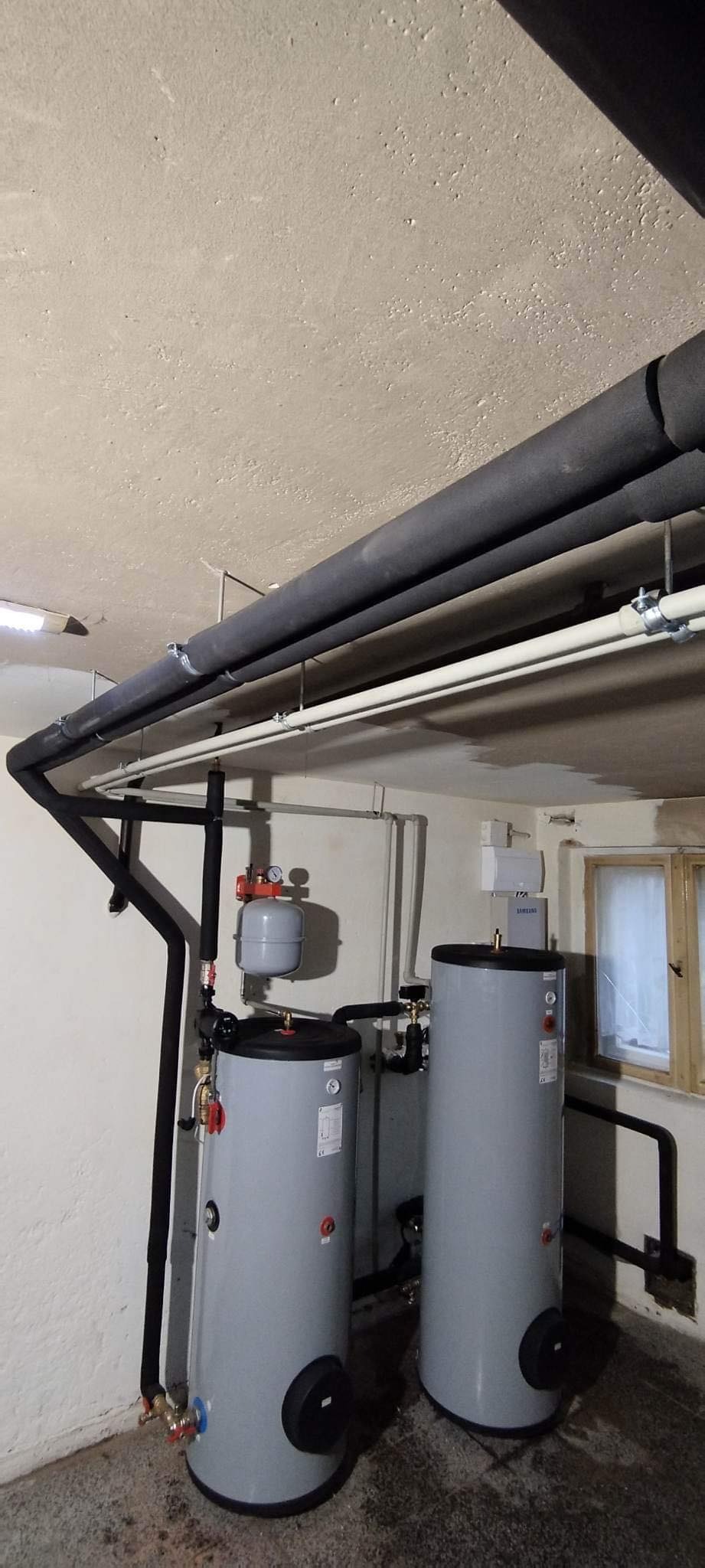 Dwa szare zbiorniki akumulacyjne ciepłej wody z czarnymi rurami izolowanymi biegnącymi wzdłuż sufitu piwnicy, widoczne okno z firanką i inne elementy instalacji.