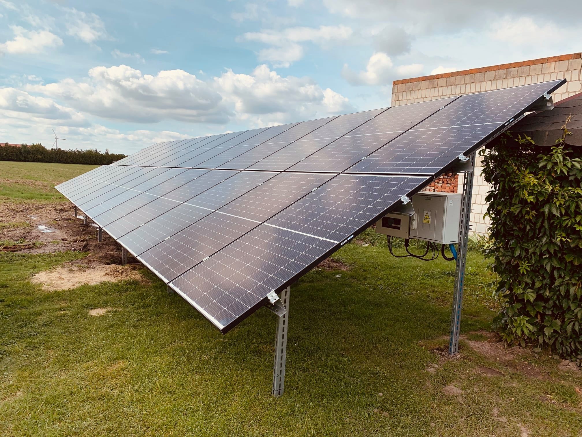 Rozległa instalacja paneli słonecznych na metalowej konstrukcji, umieszczona na trawiastym terenie z widocznym w tle wiatrakiem i budynkiem z cegły.