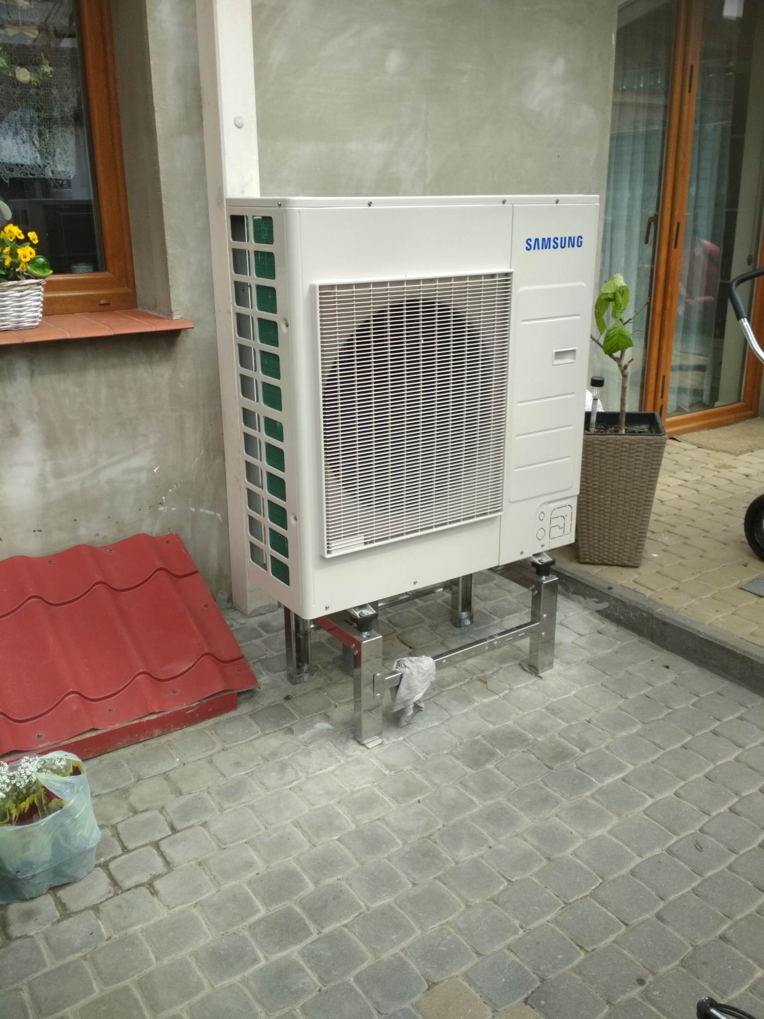 Zewnętrzna jednostka klimatyzacyjna Samsung zamontowana na metalowej konstrukcji, widoczna na tle szarej kostki brukowej i fragmentu czerwonej blachy dachowej.