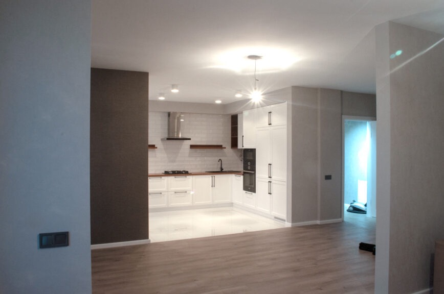 Nowoczesna kuchnia z białymi szafkami, drewnianymi blatami i płytkami metra na ścianie, widziana z perspektywy salonu z drewnianą podłogą.