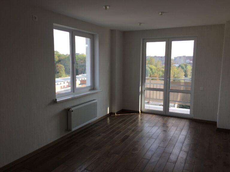 Pusty pokój z parkietem, grzejnikiem pod oknem i wyjściem na balkon. Widok z okna na drzewa i budynki.