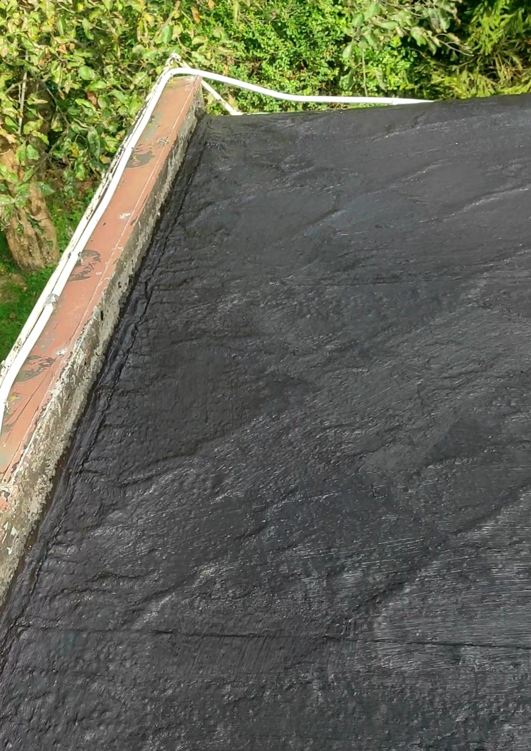 Widok z góry na fragment pokrycia dachu z papy bitumicznej po renowacji, z widocznymi nierównościami i teksturą, obok krawędzi dachu z brązowym obrzeżem i fragmentami roślinności w tle.