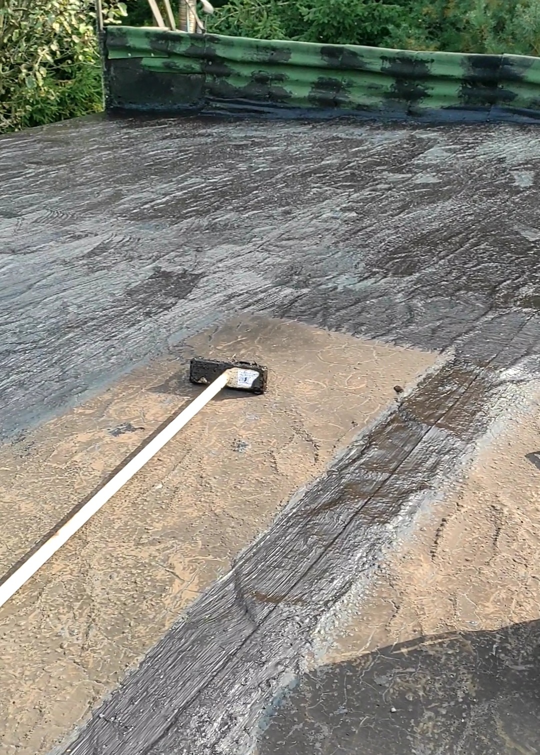 Hydroizolacja dachu płaskiego smołą za pomocą szczotki na długim kiju; widoczny fragment dachu po renowacji, zieleń w tle.