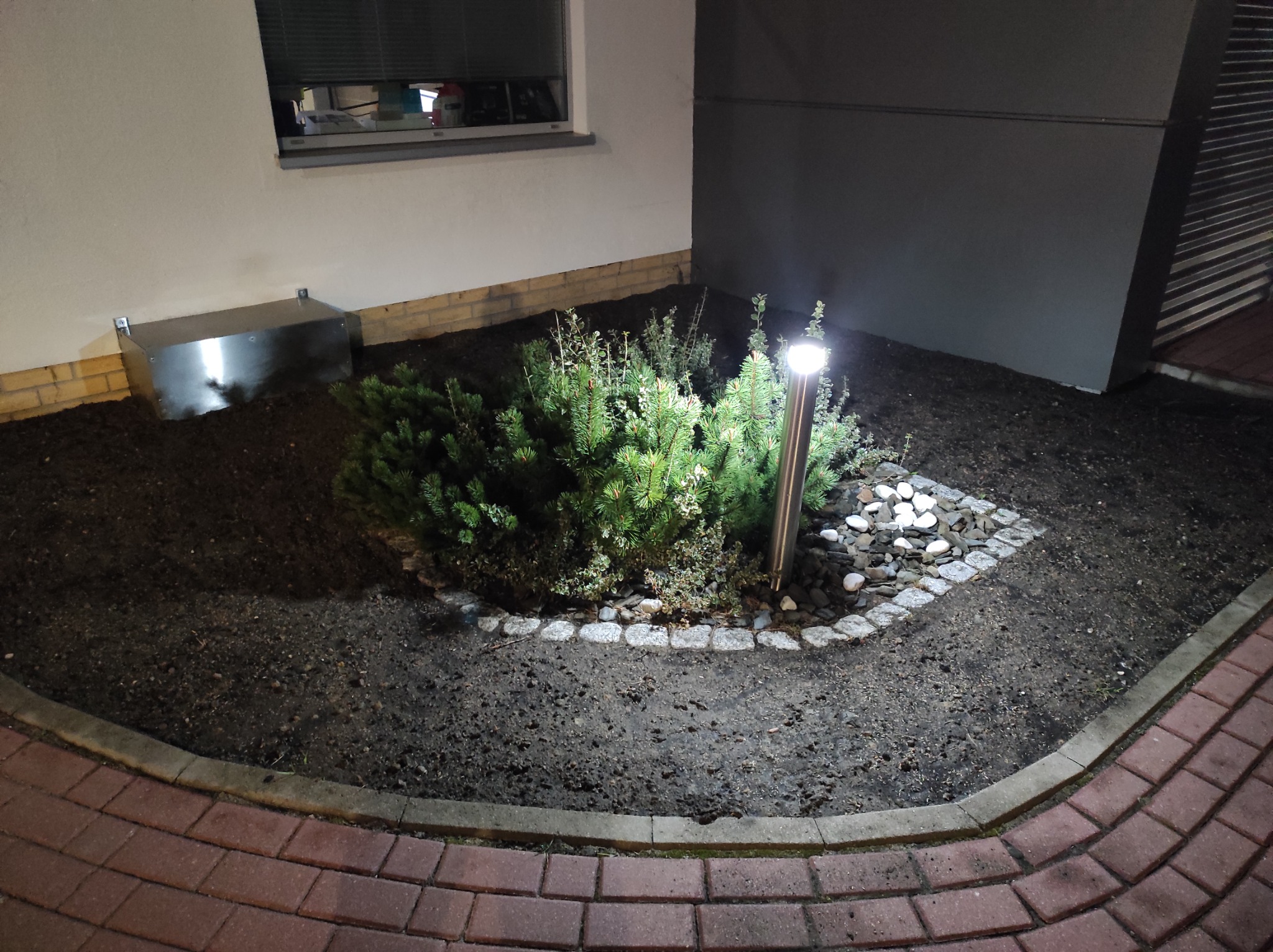 Niewielka rabata z iglakami i ozdobnymi kamieniami, oświetlona lampą ogrodową, na tle elewacji budynku.