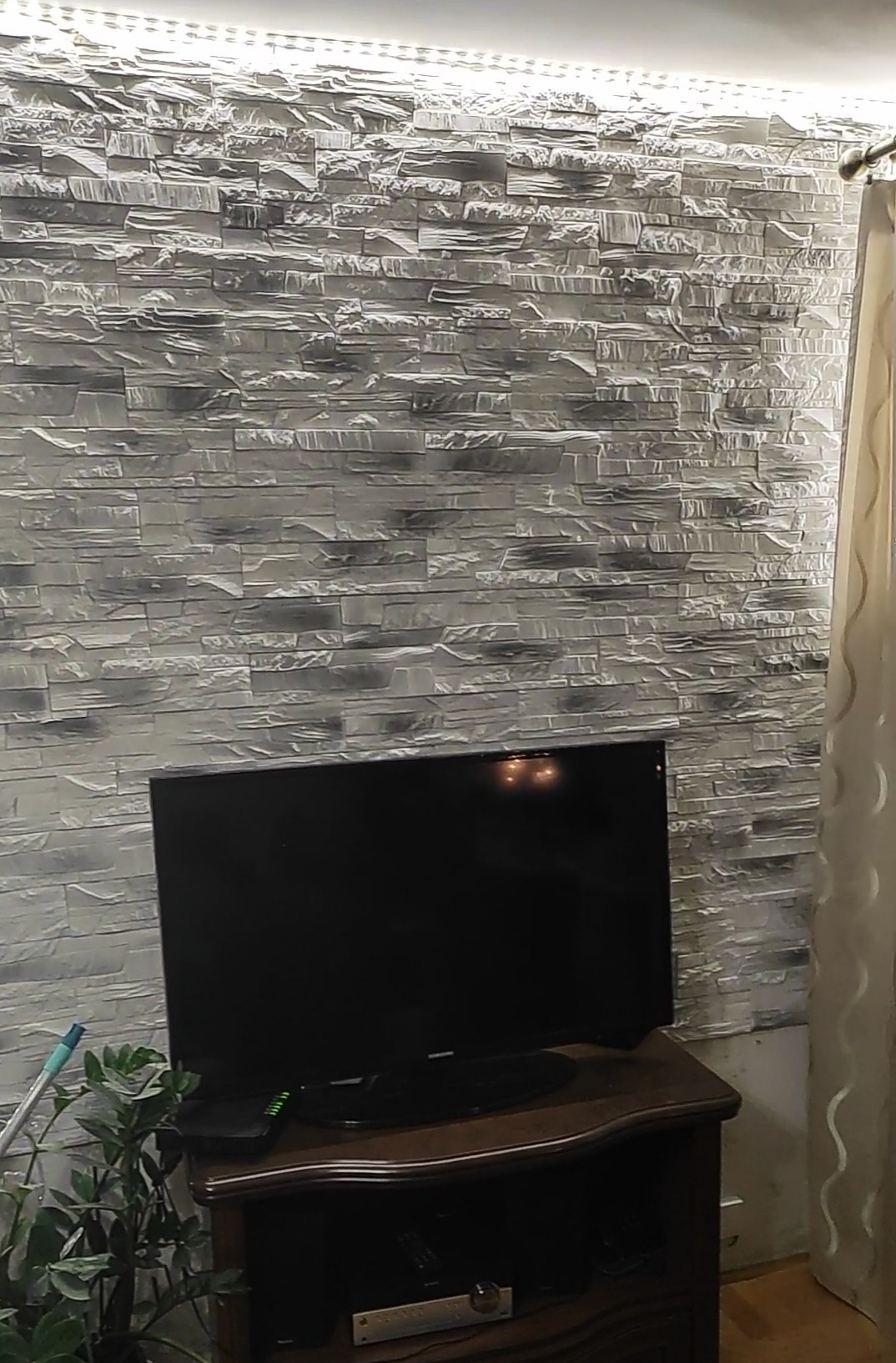 Ściana z dekoracyjnej, szarej cegły z podświetleniem LED u góry, telewizor na drewnianej komodzie, zasłona w tle i roślina doniczkowa po lewej stronie.