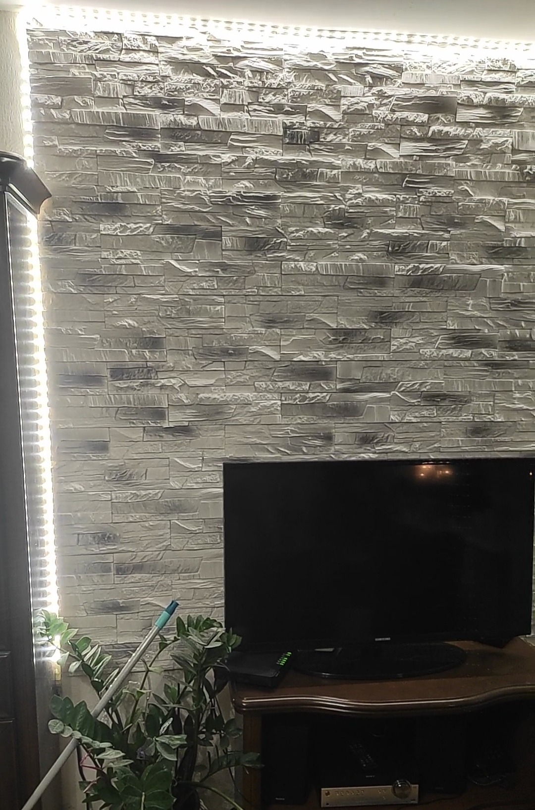Dekoracyjna ściana z imitacji kamienia z podświetleniem LED w Gdańsku, obok telewizor i roślina doniczkowa.