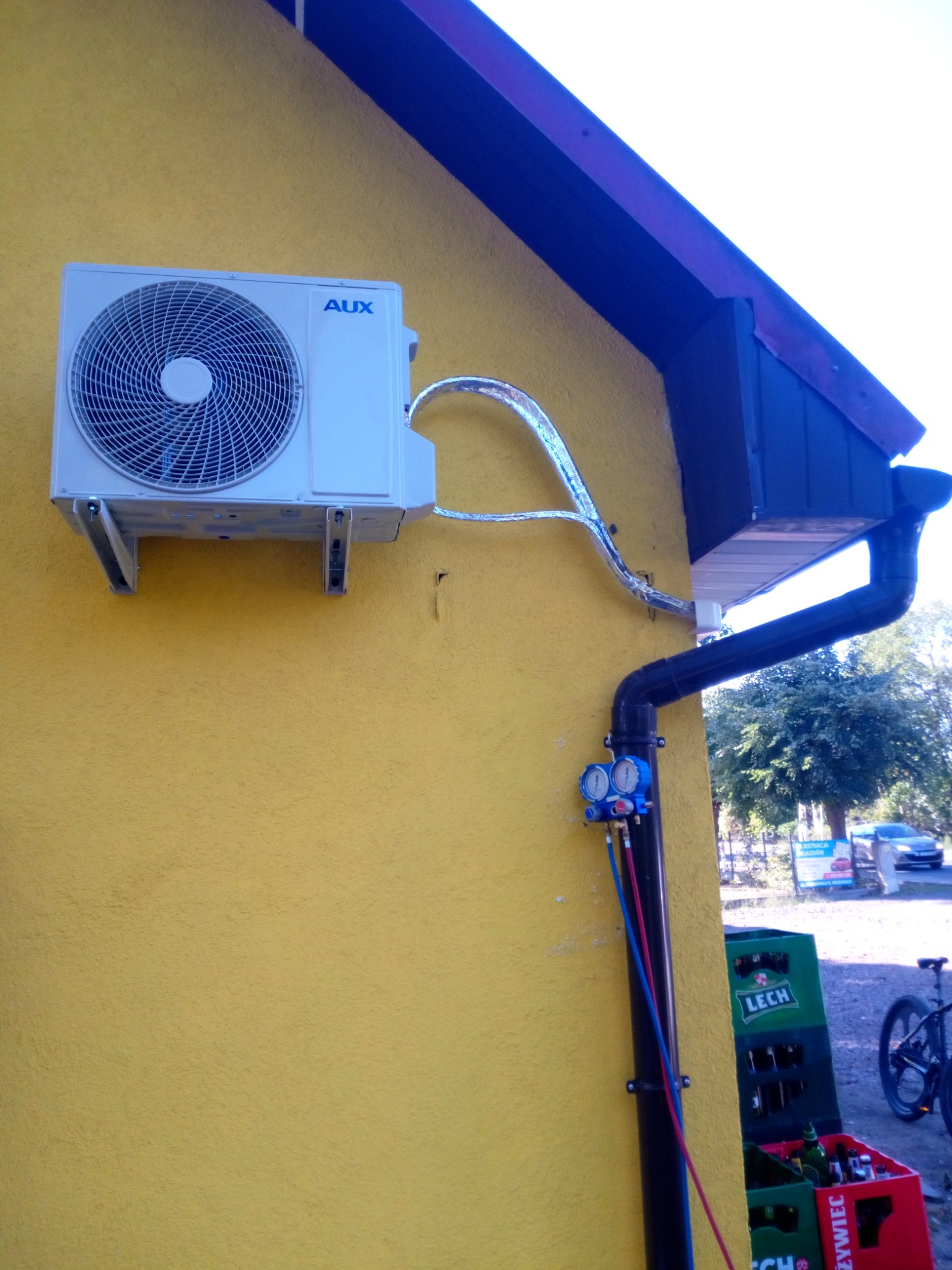 Zamontowana jednostka zewnętrzna klimatyzacji AUX na żółtej ścianie budynku, z widocznymi przewodami i manometrami serwisowymi. W tle widoczne skrzynki z butelkami i rower.