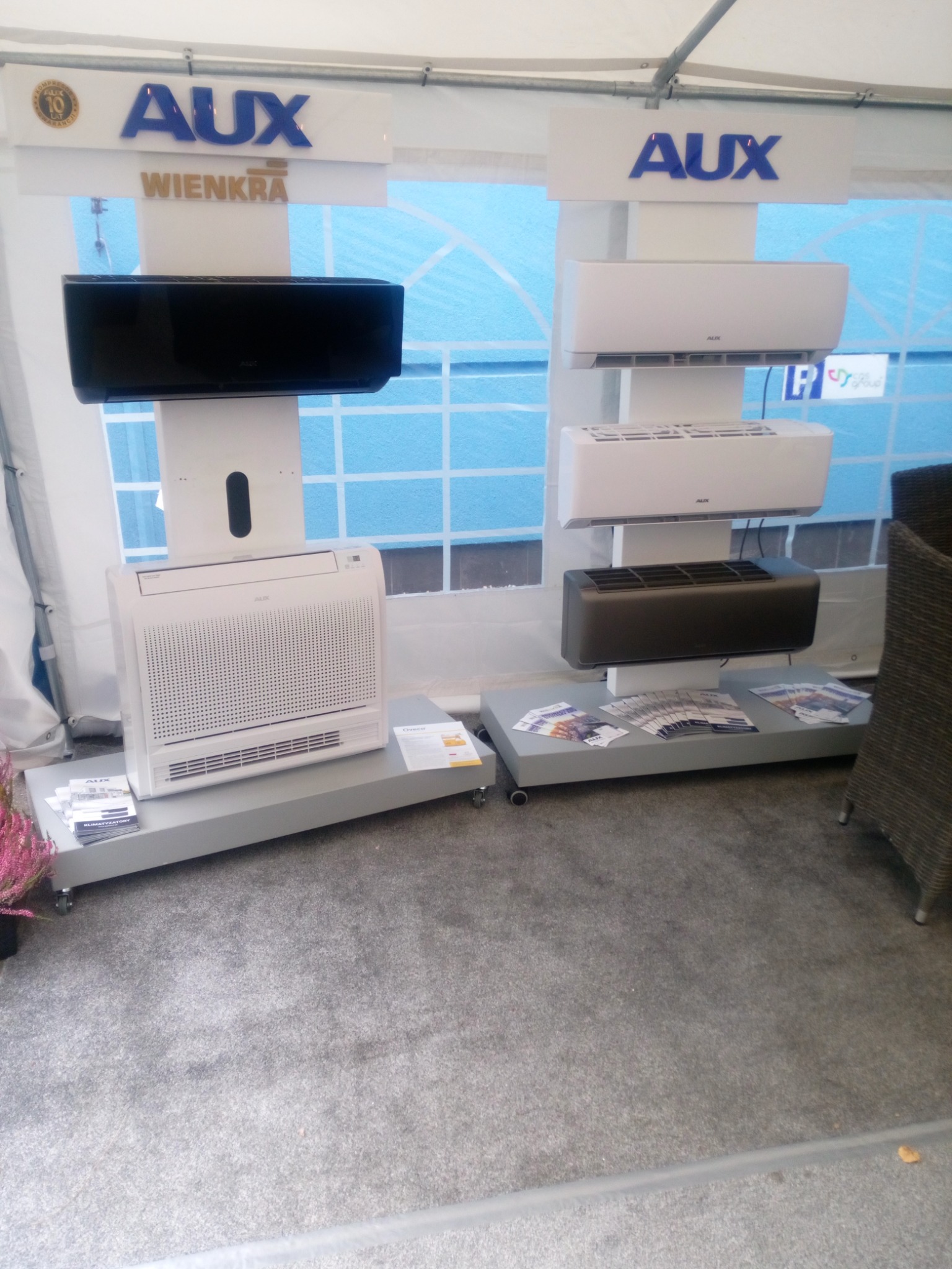 Ekspozycja różnych modeli klimatyzatorów marki AUX na szarym dywanie, widoczne modele ścienne w kolorze białym i czarnym oraz model podłogowy, ulotki informacyjne na podeście.