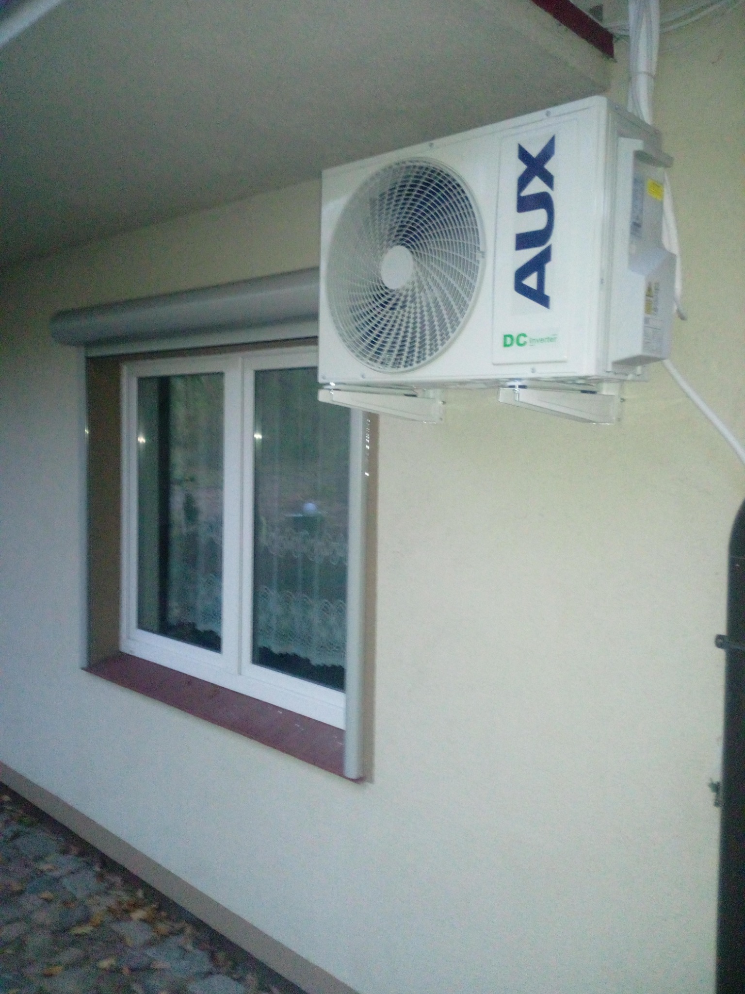 Zewnętrzna jednostka klimatyzacji AUX DC Inverter zamontowana na ścianie budynku obok okna z roletą zewnętrzną.