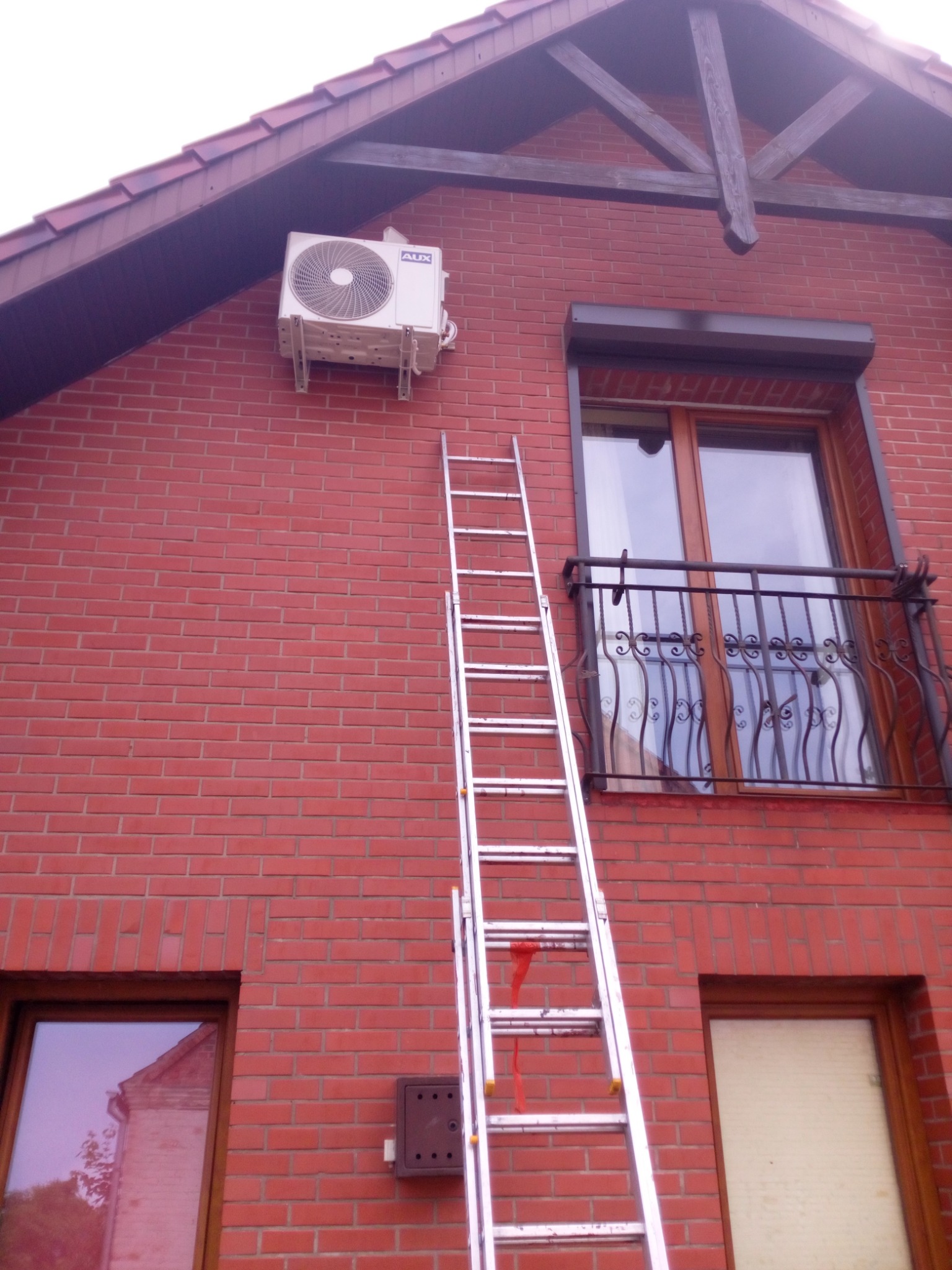 Zewnętrzna jednostka klimatyzacji marki AUX zamontowana na czerwonej ceglanej ścianie domu jednorodzinnego, z drabiną opartą o ścianę, prowadzącą do urządzenia.
