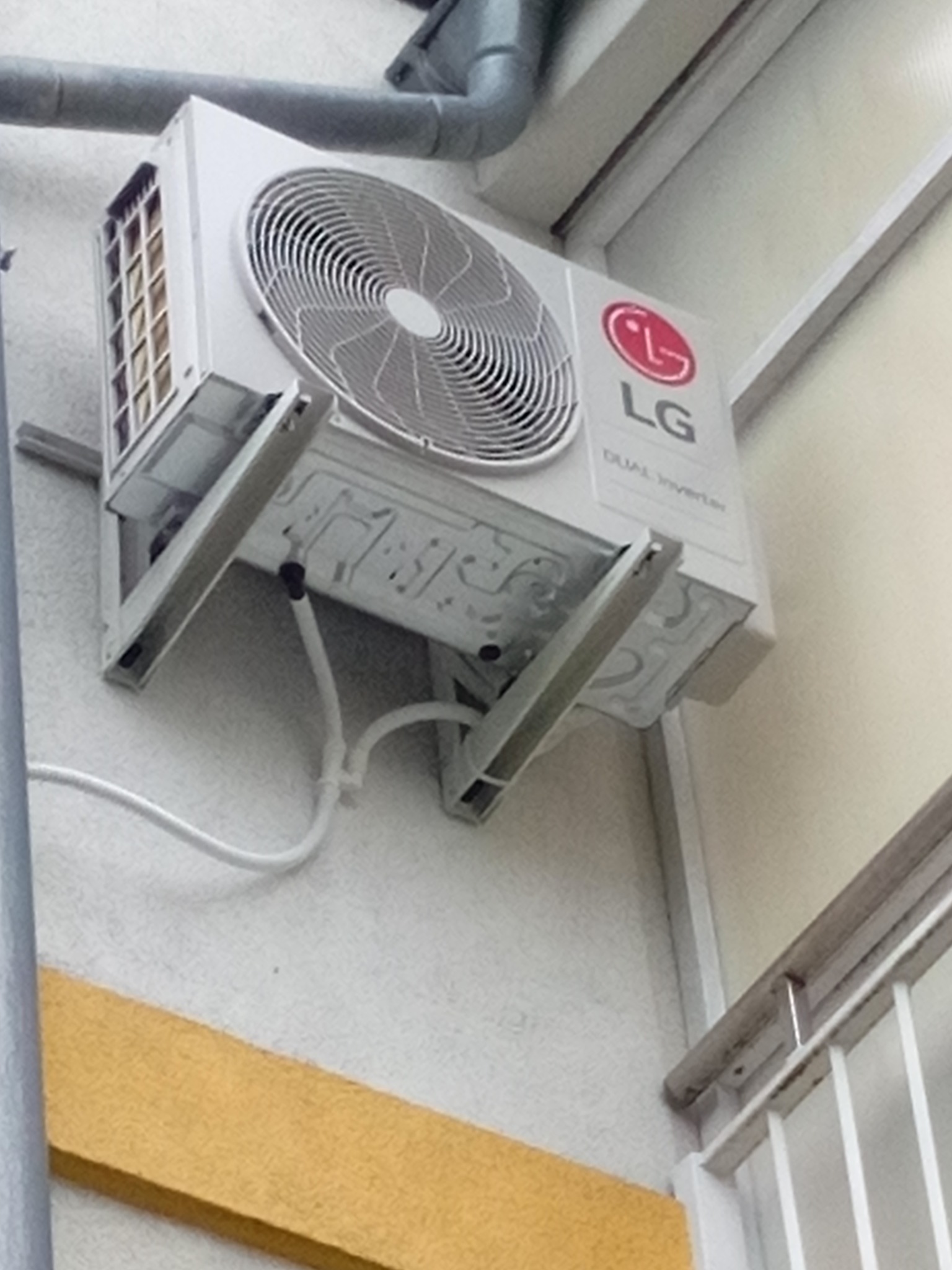 Zewnętrzna jednostka klimatyzacji LG Dual Inverter zamontowana na wspornikach na elewacji budynku, widoczne rury odprowadzające skropliny, ujęcie z dołu.
