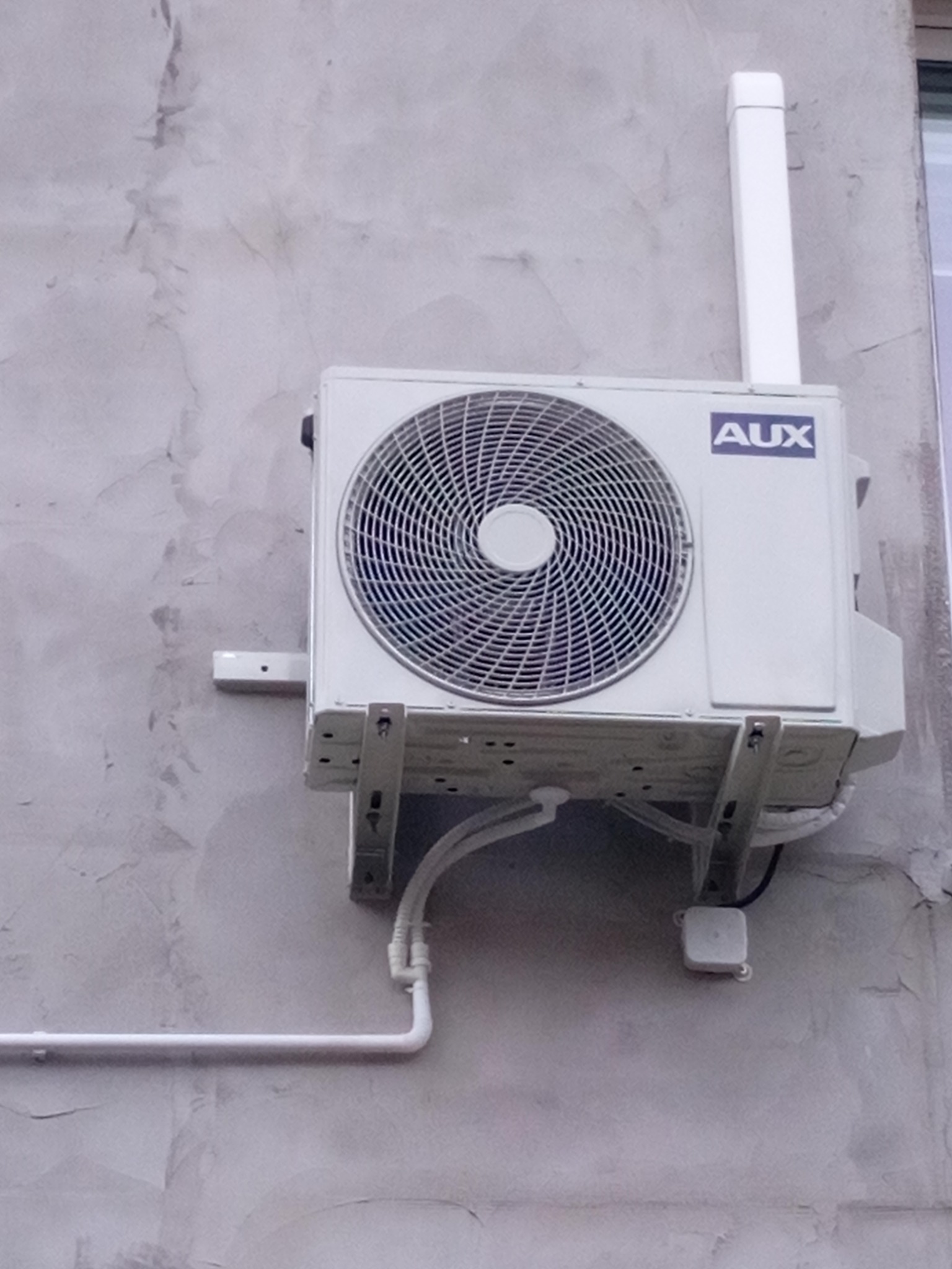 Zewnętrzna jednostka klimatyzacji marki AUX zamontowana na szarej, chropowatej ścianie budynku, widoczne rury odprowadzające skropliny.