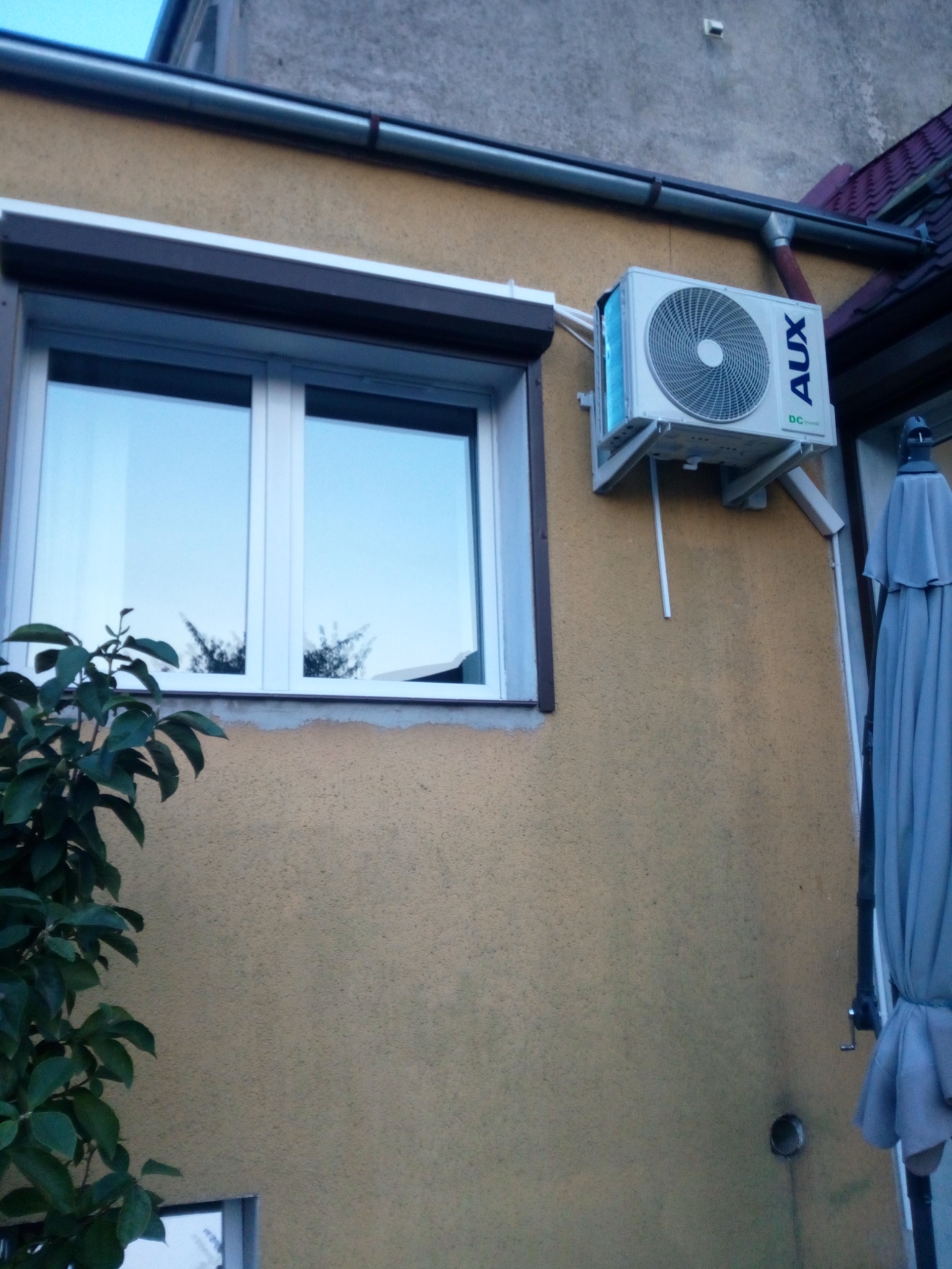 Zewnętrzna jednostka klimatyzacji AUX, zamontowana na elewacji budynku obok okna z roletą, widoczny fragment parasola ogrodowego.