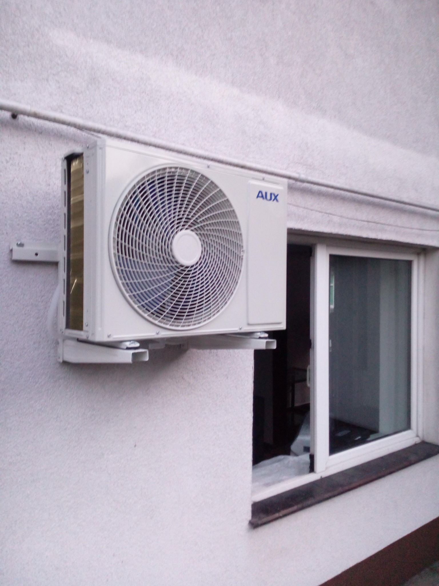 Zewnętrzna jednostka klimatyzacji marki AUX zamontowana na ścianie budynku obok okna. Widoczne uchwyty mocujące i instalacja odprowadzająca skropliny.