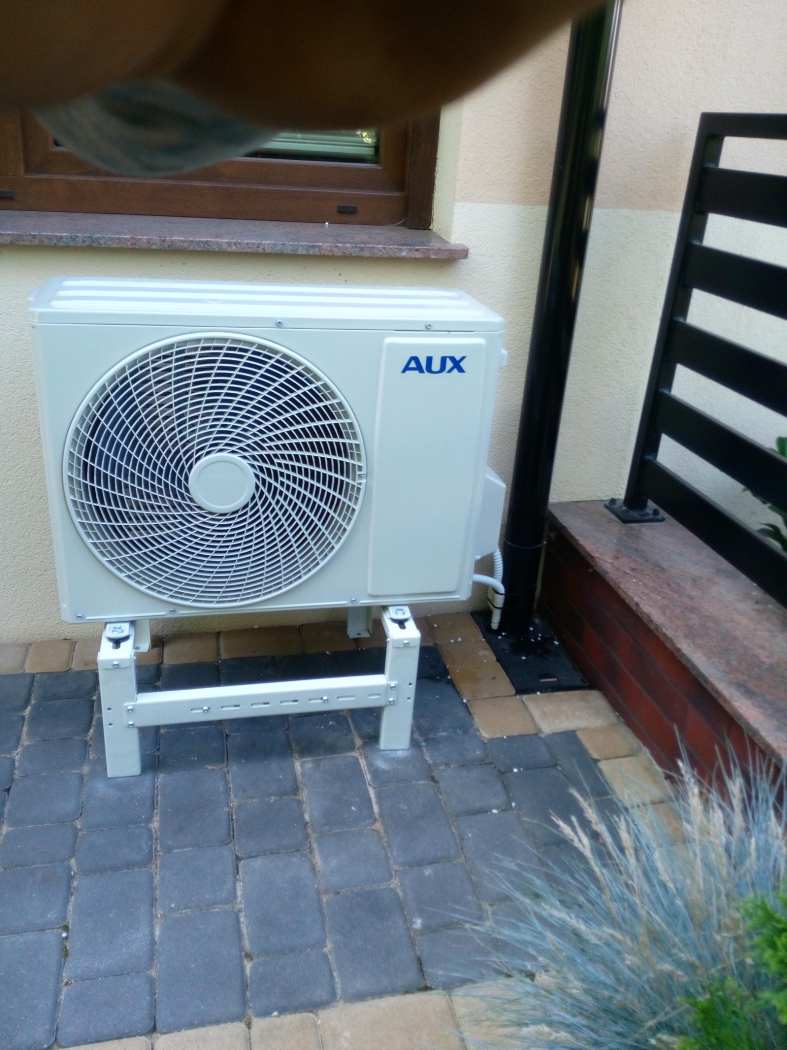 Zewnętrzna jednostka klimatyzacji marki AUX, zamontowana na metalowym stelażu na kostce brukowej przed budynkiem. Widoczna rura odprowadzająca skropliny i fragment okna z kamiennym parapetem.