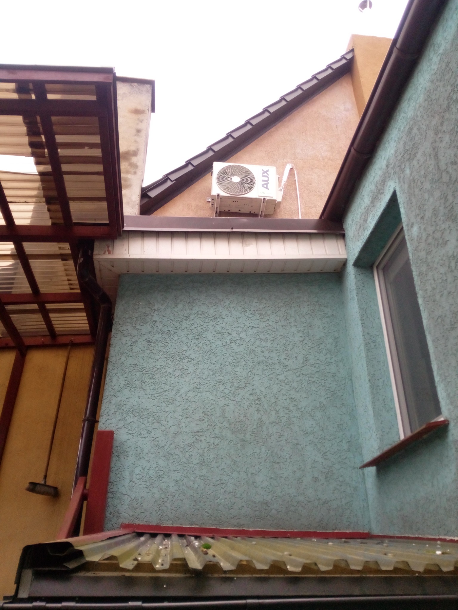 Zewnętrzna jednostka klimatyzacji AUX zamontowana na elewacji domu, widoczna z dołu, z fragmentami dachu i rynny w kadrze.
