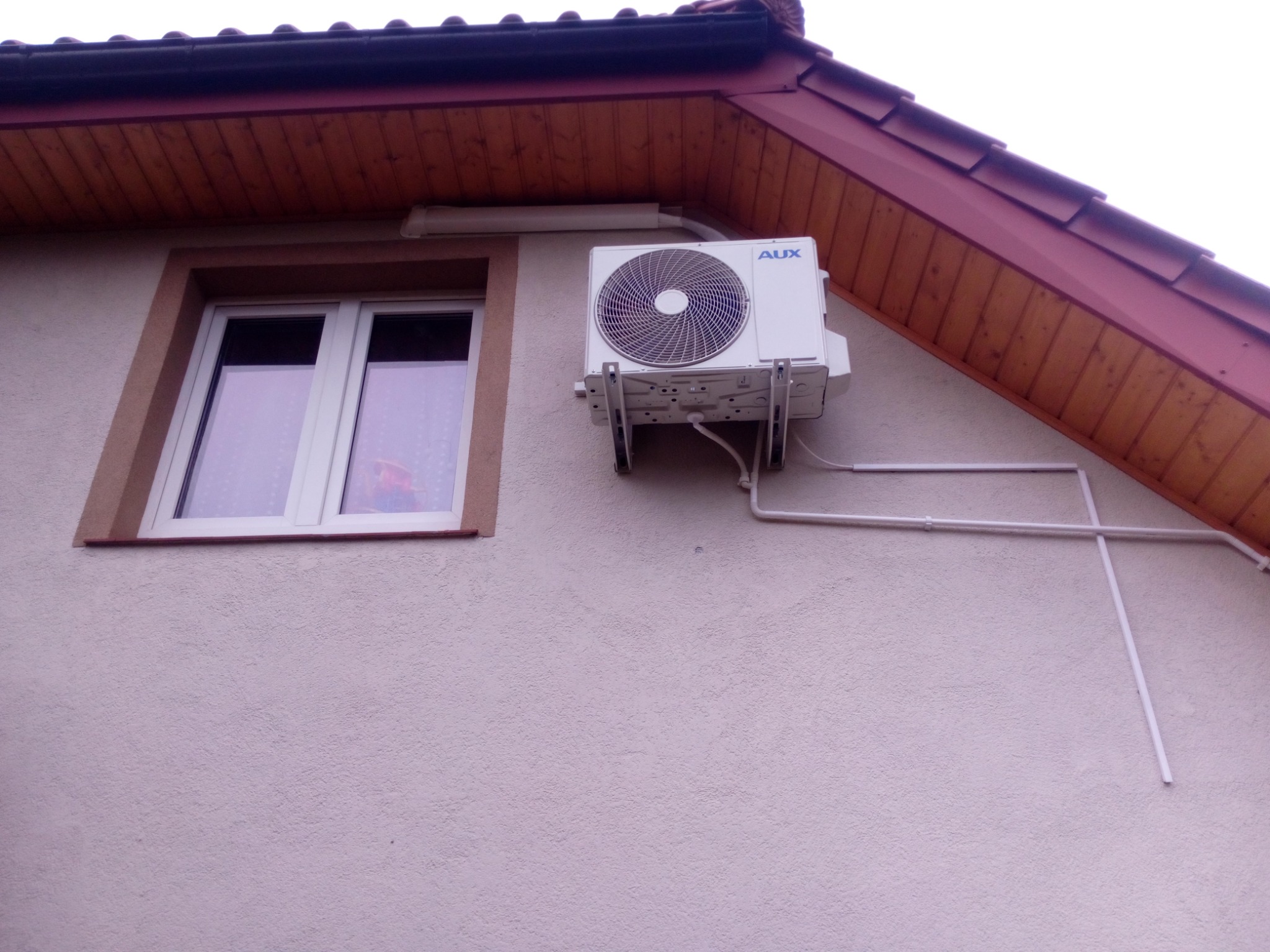 Biała jednostka zewnętrzna klimatyzatora marki AUX zamontowana na elewacji domu z oknem, widoczne rury odprowadzające skropliny.