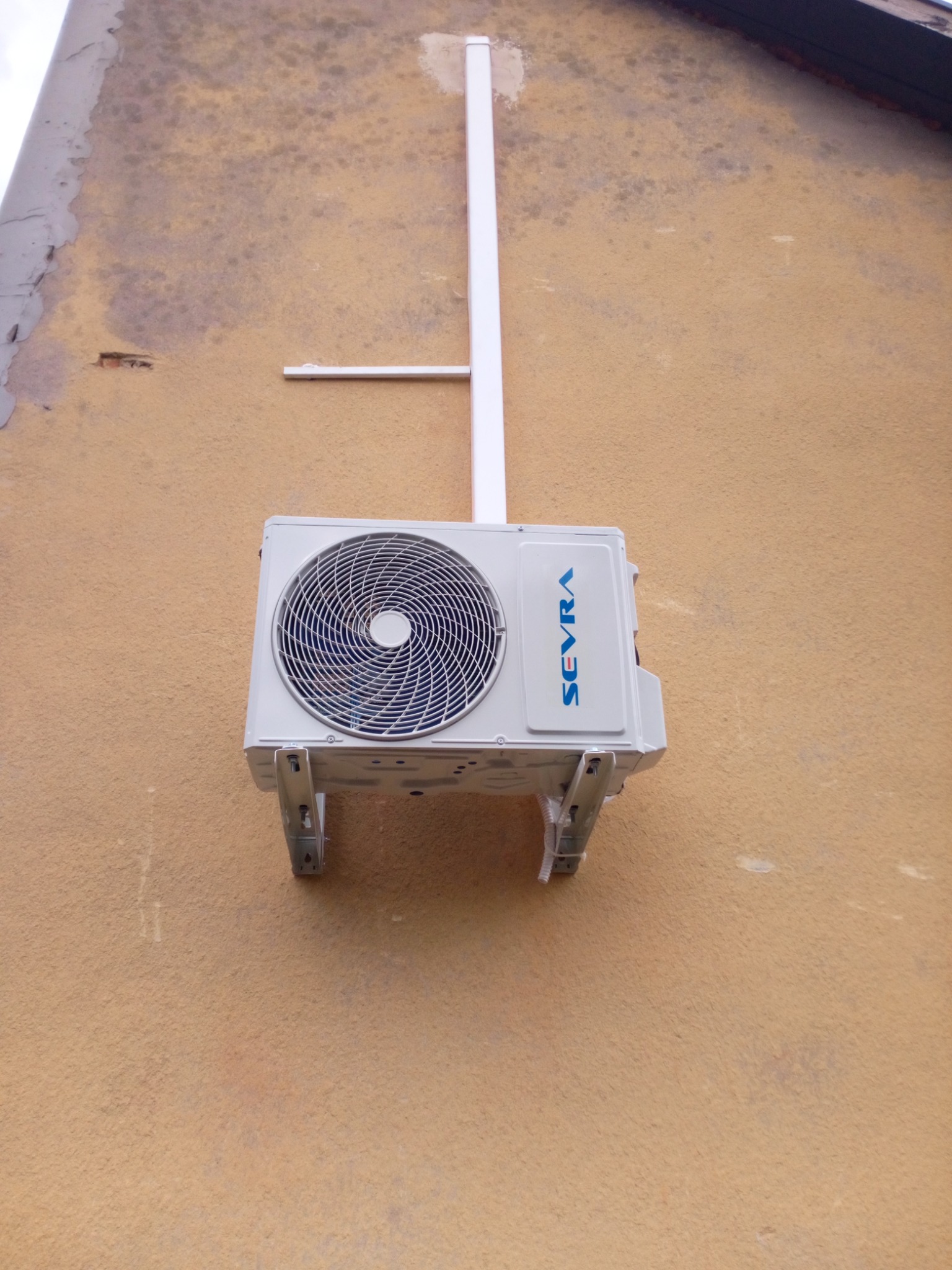 Zewnętrzna jednostka klimatyzatora marki Sevra, zamontowana na wspornikach na elewacji budynku o pomarańczowej, lekko zabrudzonej ścianie. Widoczna instalacja odprowadzająca skropliny.