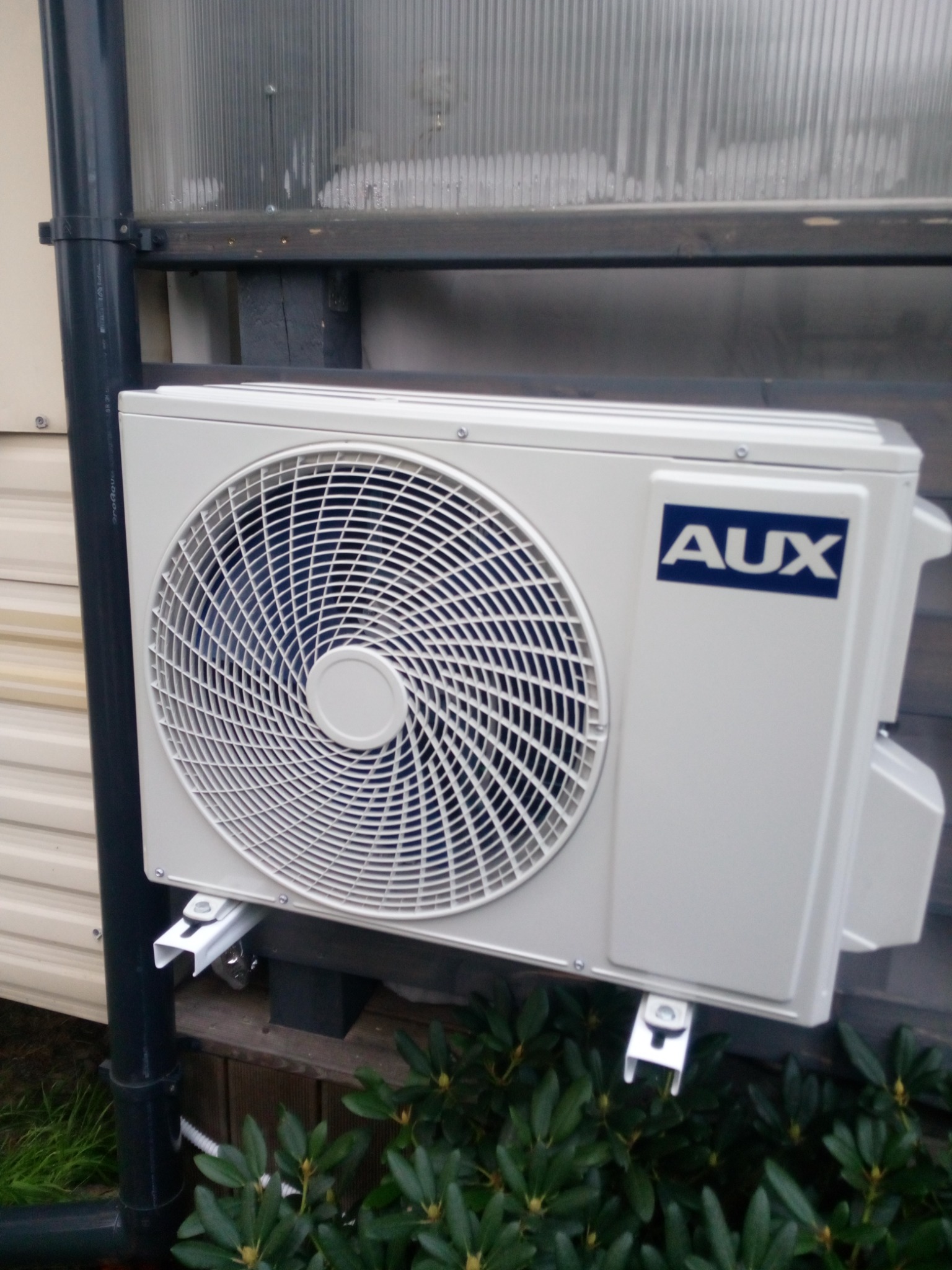Zewnętrzna jednostka klimatyzacji marki AUX, zamontowana na drewnianej elewacji budynku, widoczna instalacja odprowadzająca skropliny, otoczona zielenią.