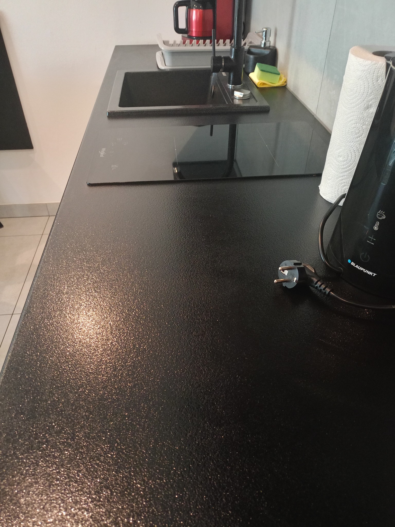 Błyszczący, czarny blat kuchenny ze zlewem, płytą indukcyjną i czajnikiem Blaupunkt. Widoczny kabel z wtyczką i rolka ręcznika papierowego.