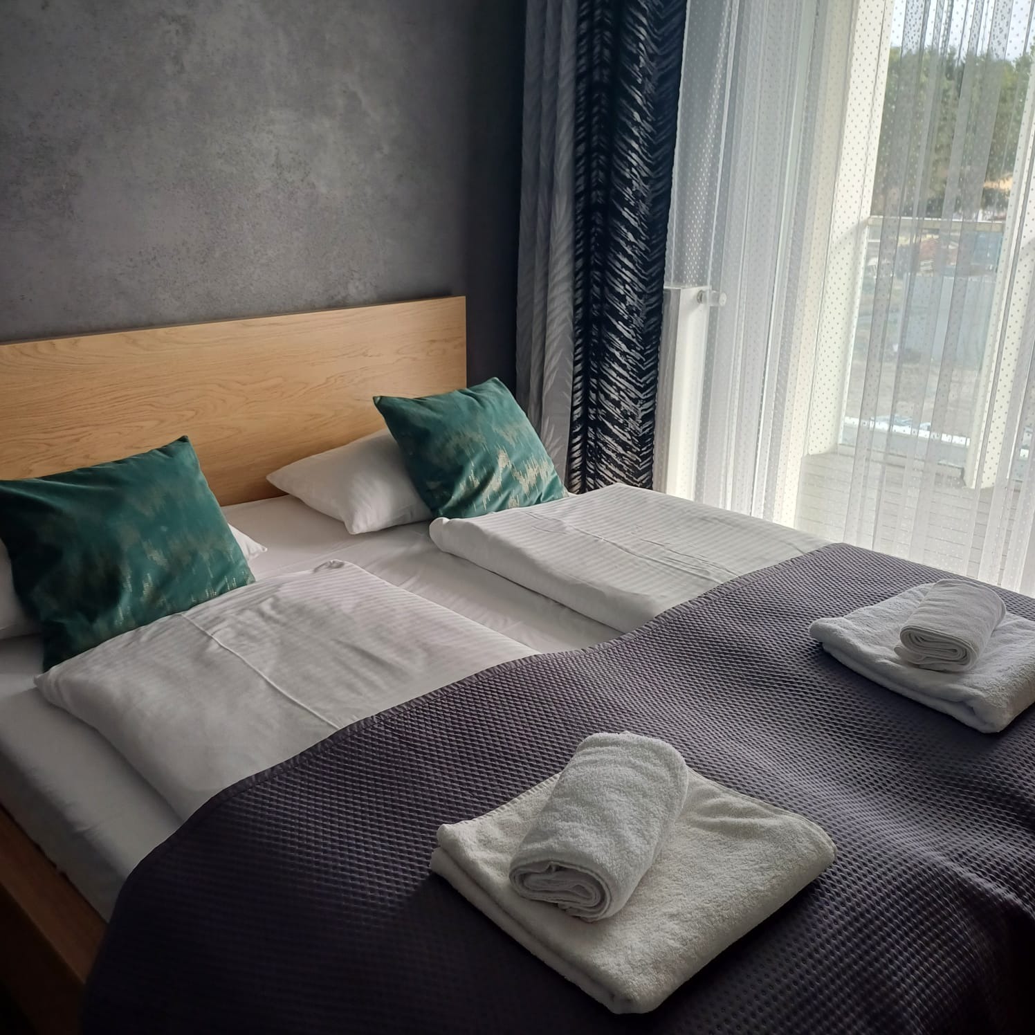 Pokój hotelowy z dwoma łóżkami, świeżo pościelony, z ręcznikami ułożonymi na kołdrze. Widoczne okno z firankami i zasłonami. Ściana w tle o fakturze betonu.