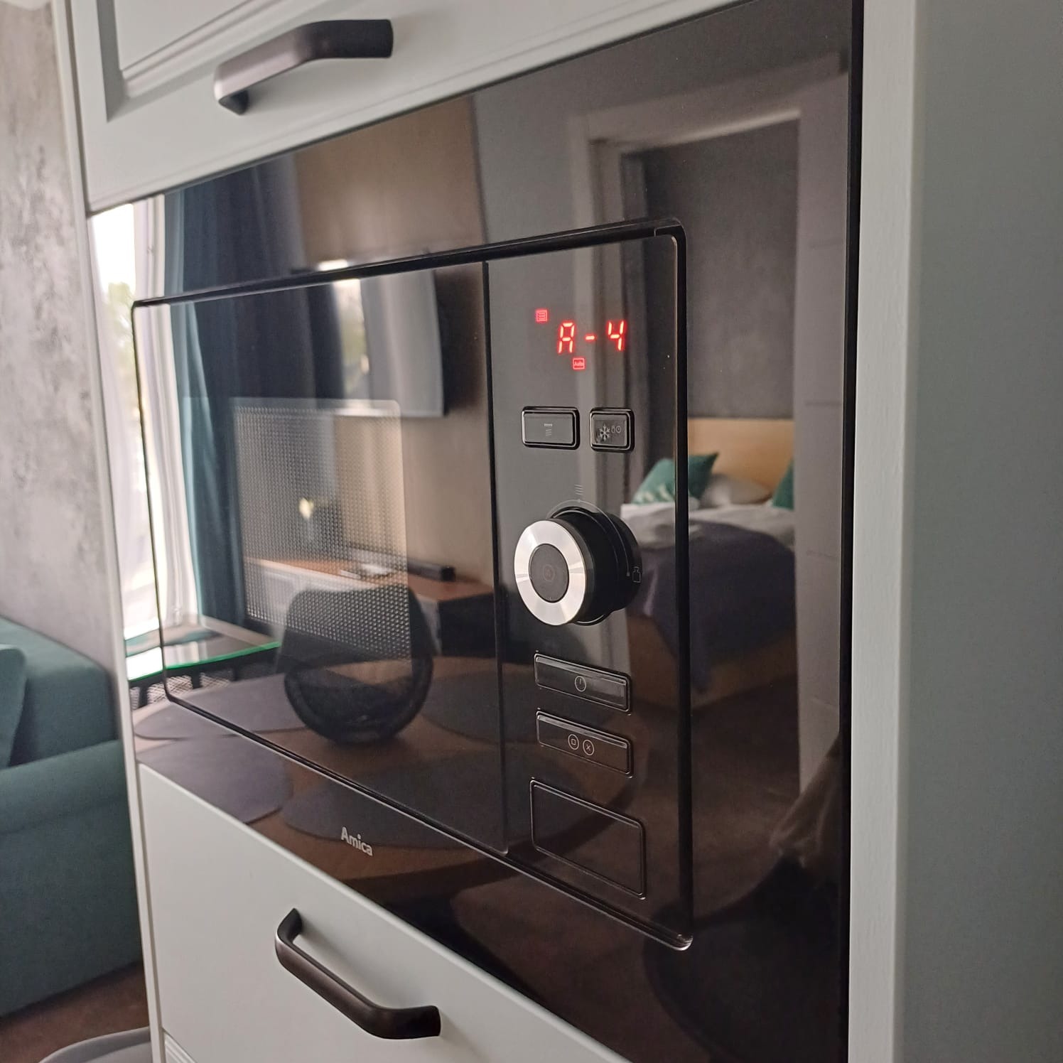 Wbudowana czarna mikrofalówka marki Amica w jasnej zabudowie kuchennej. Widoczne odbicie wnętrza pokoju w szklanych drzwiach urządzenia.