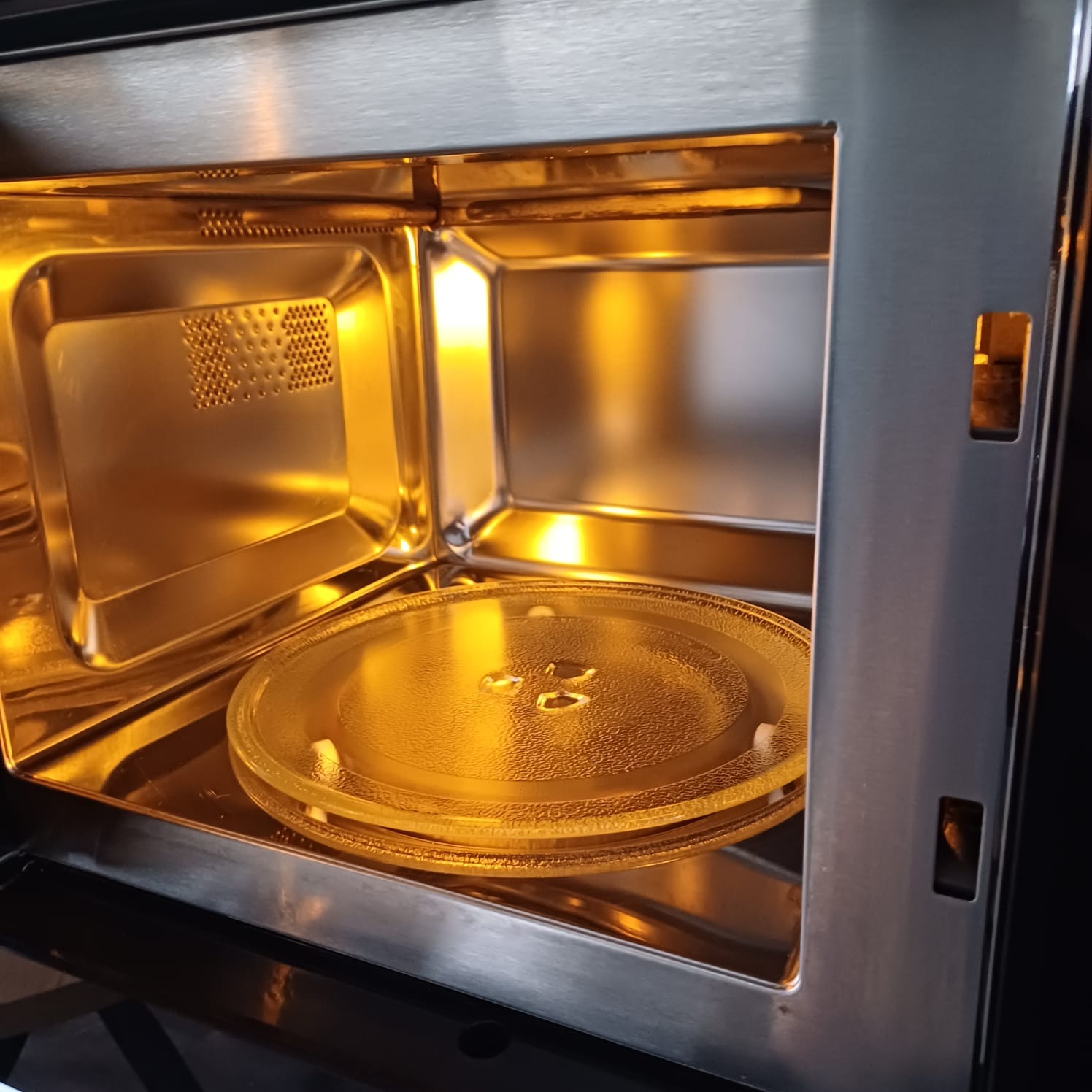 Wnętrze czystej mikrofalówki ze szklanym talerzem obrotowym, oświetlone ciepłym, żółtym światłem. Metalowe ścianki i perforowany panel wentylacyjny.