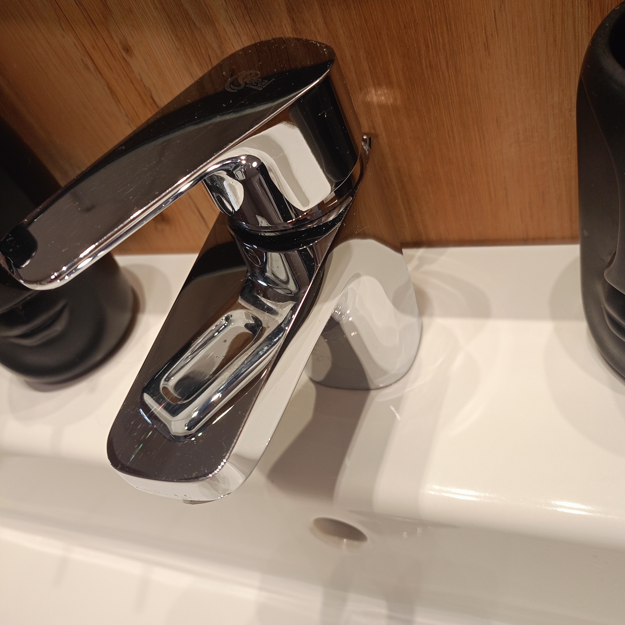 Błyszcząca, chromowana bateria umywalkowa w minimalistycznym stylu, zamontowana na białej umywalce z drewnianym panelem w tle.