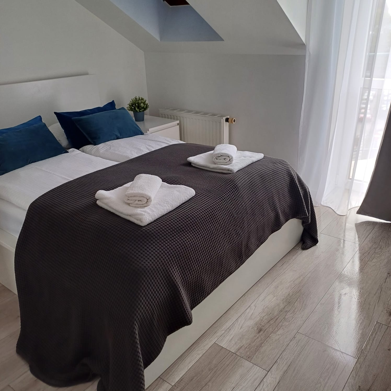 Jasna sypialnia na poddaszu z drewnianą podłogą. Na łóżku z ciemnym pledem leżą białe, złożone ręczniki i niebieskie poduszki. Białe zasłony wpadają do pokoju przez balkon.