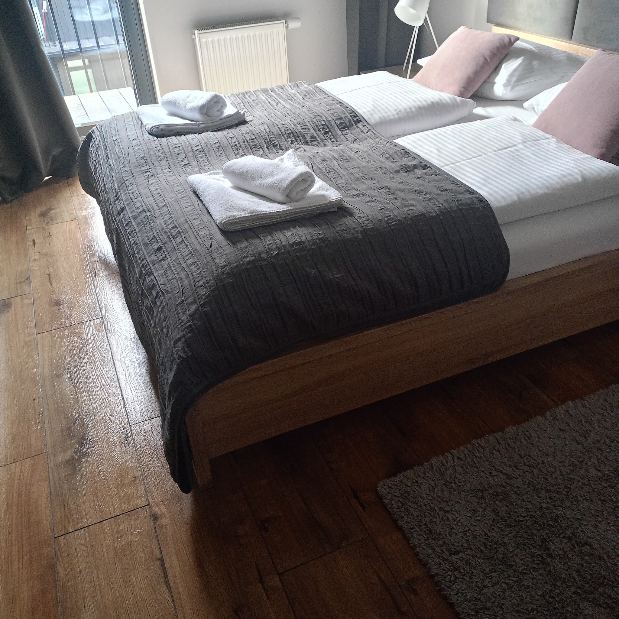 Pokój hotelowy z dwuosobowym łóżkiem, na którym leżą białe ręczniki. Na podłodze widoczny dywanik. Za oknem balkon.