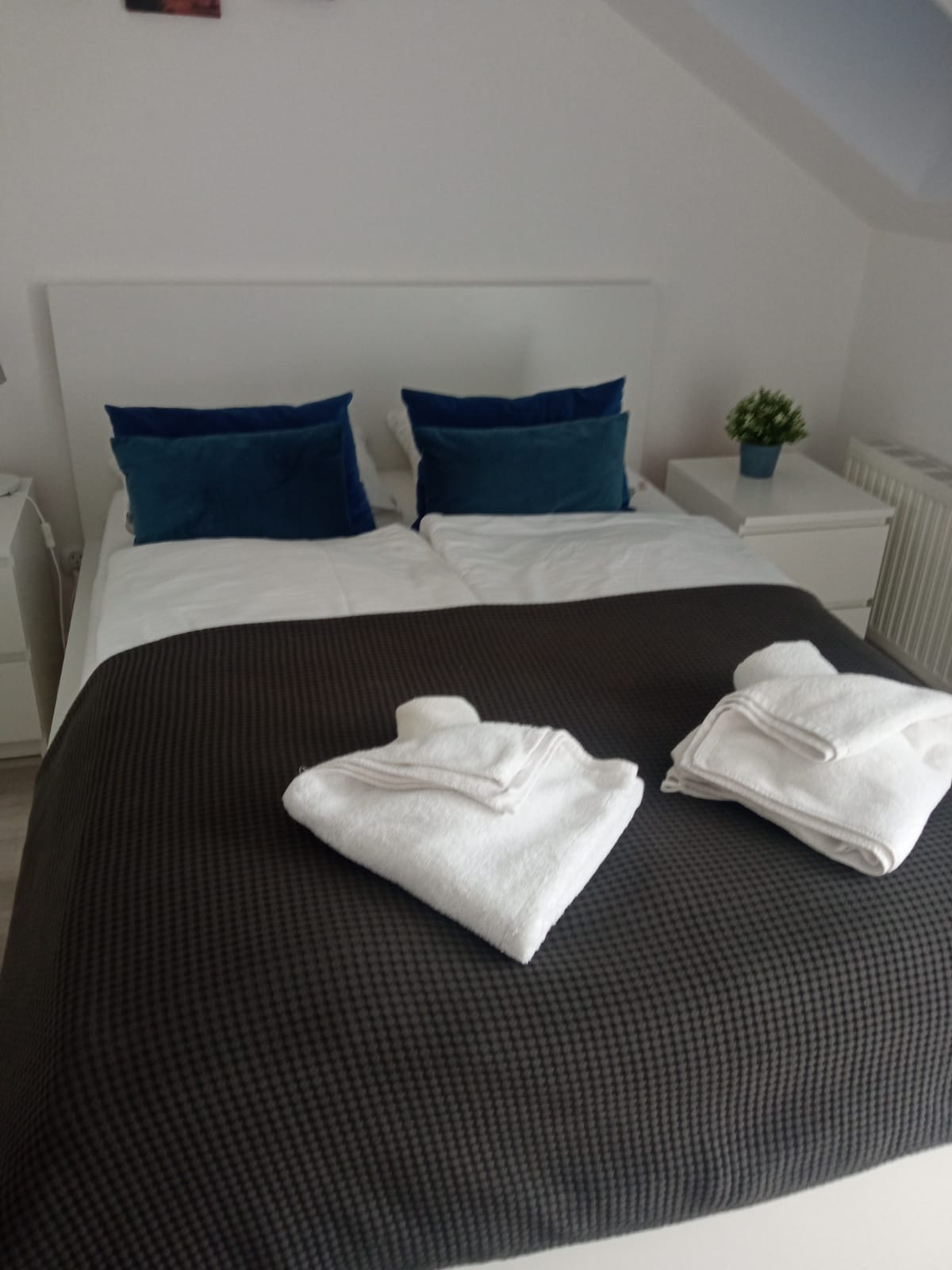 Czysta pościel na łóżku z ciemnym pledem i złożonymi ręcznikami w schludnym pokoju z białymi ścianami i szafkami nocnymi. Roślina w doniczce na szafce nocnej.