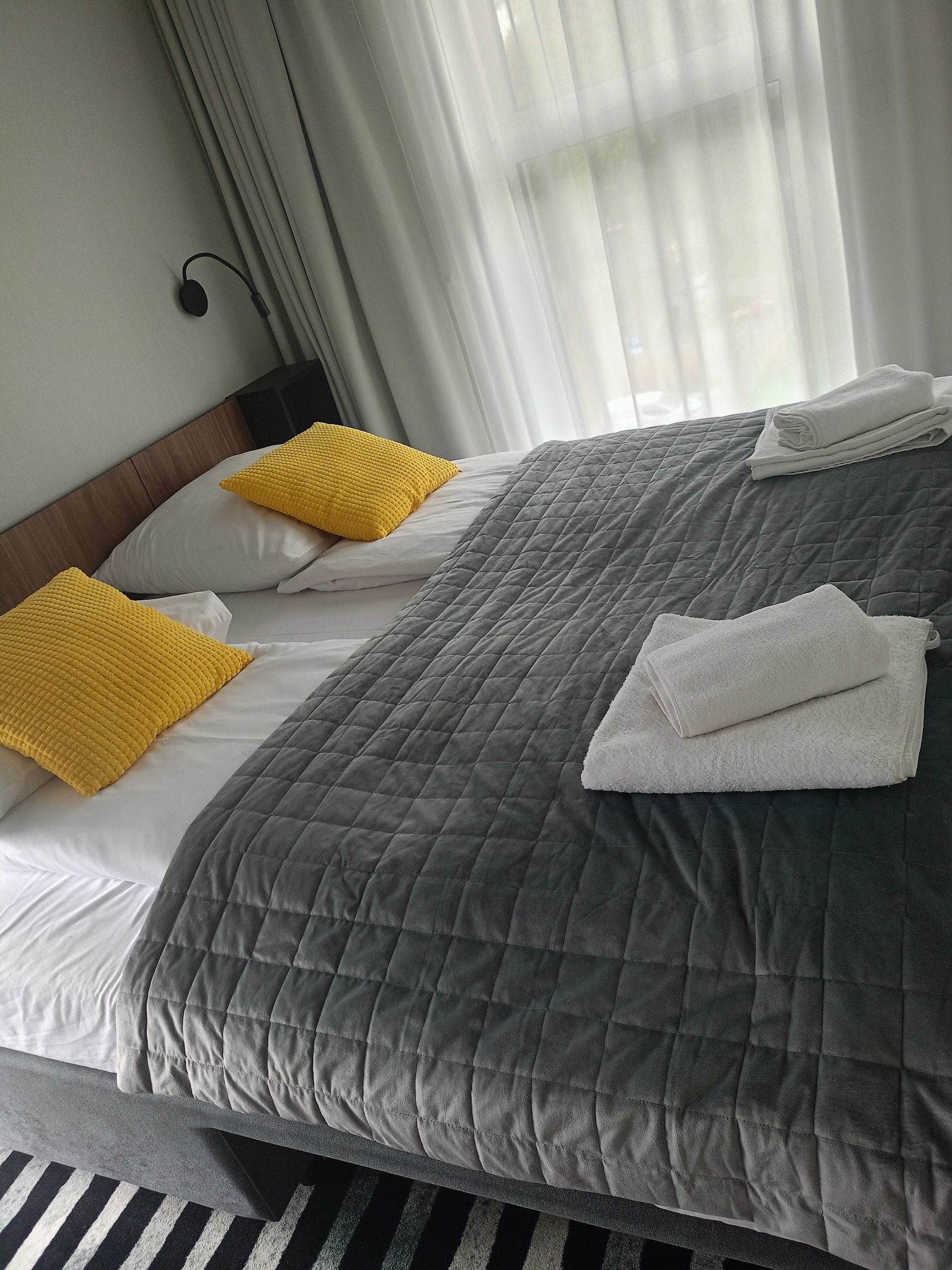 Wnętrze pokoju hotelowego: starannie pościelone łóżko z szarą narzutą w kratę, żółtymi poduszkami i stosem białych ręczników, oświetlone naturalnym światłem z okna.