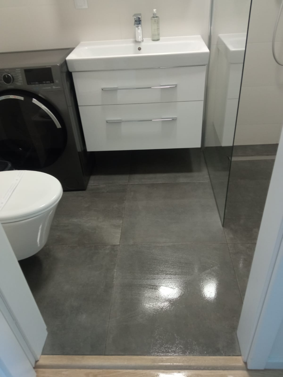 Umywalka z szafką, pralka i toaleta w nowoczesnej łazience z szarymi płytkami podłogowymi. Widoczna kabina prysznicowa ze szklaną ścianą.