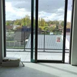 Wnętrze z dużym przesuwnym oknem balkonowym, widok na zewnątrz z metalową balustradą i dachami domów, na podłodze ułożone jasne płytki, w rogu paczka płytek.