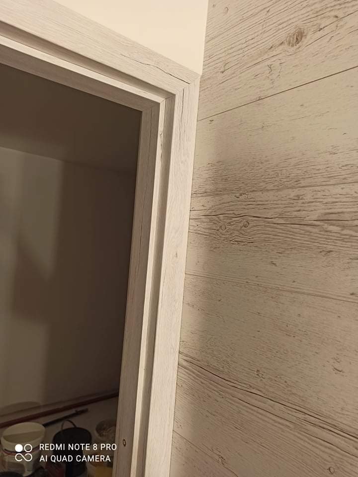 Ściana wykończona panelami imitującymi drewno w odcieniach bieli i szarości, z widoczną ościeżnicą drzwiową w podobnej kolorystyce, ujęcie z dołu, ukazujące fakturę materiałów.