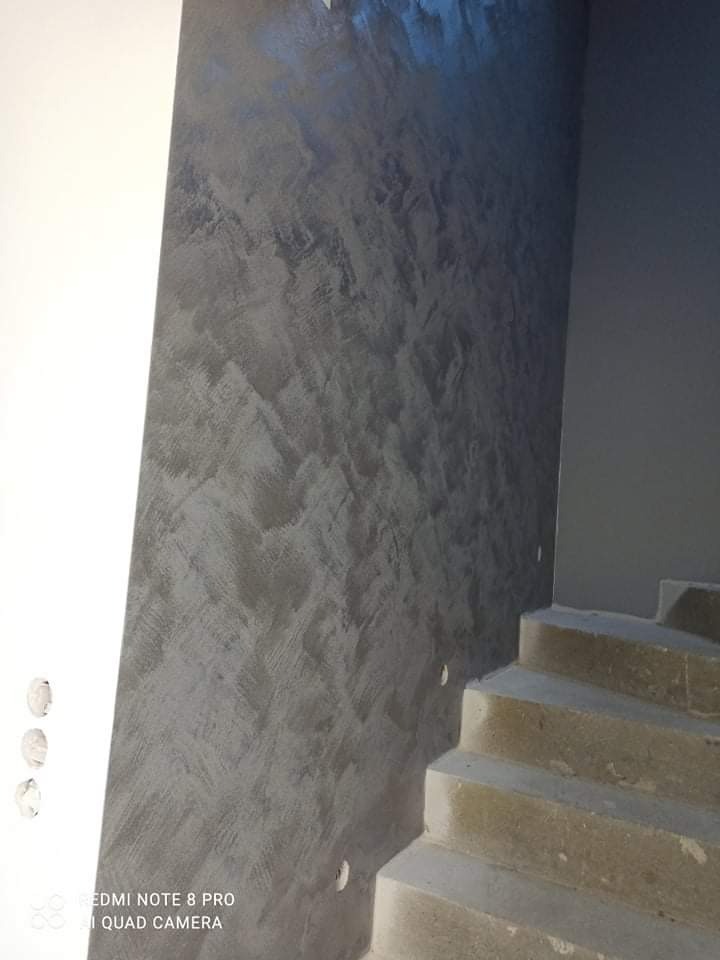 Ściana wewnętrzna z dekoracyjnym, szarym tynkiem strukturalnym, widoczne fragmenty schodów w surowym stanie.